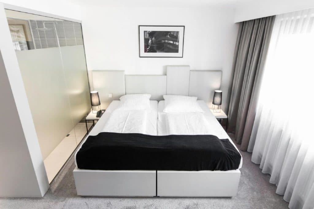 Minimalistisches Hotelzimmer mit weißem Doppelbett, schwarzem Läufer, Nachttischlampen und einer mattierten Glastrennwand.